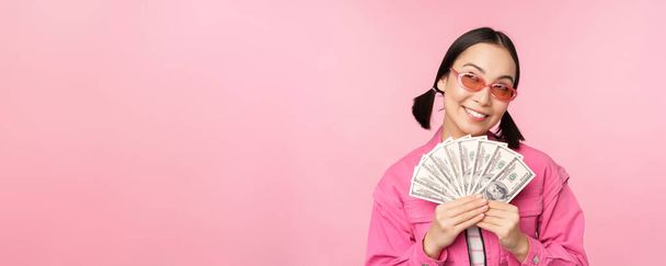 Красива корейська жінка в сонцезахисних окулярах, показує долари, готівку, усміхнені задоволені, концепція швидких кредитів, мікрокредитування та оплати, стоїть на рожевому тлі
 - Фото, зображення