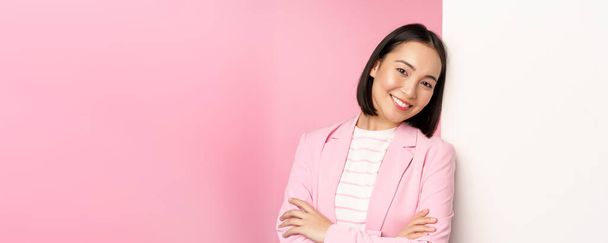 Itsevarma, menestyvä japanilainen toimisto nainen puku, rajat aseita, etsivät kuin ammattilainen kamera, nojaten valkoinen seinä mainos, tyhjä kopio tilaa logo, vaaleanpunainen tausta - Valokuva, kuva