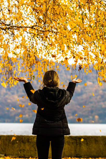 Back view of alone woman enjoying autumn, throwing fallen leaves on autumn alley. Autumn landscape, orange foliage in a park in Orsova, Romania, 2020 - Photo, Image