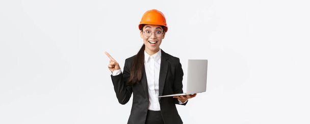 Nadšený asijský ženský inženýr, průmyslová žena v bezpečnostní přilbě a obleku, ukazující prezentaci, ukazující prstem na graf nebo graf a držící notebook, usmívající se - Fotografie, Obrázek