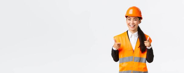 Confident pretty asian female construction manager in safety helmet and reflective jacket, pointing fingers at camera praising nice choice, good work or well done, standing white background - Zdjęcie, obraz
