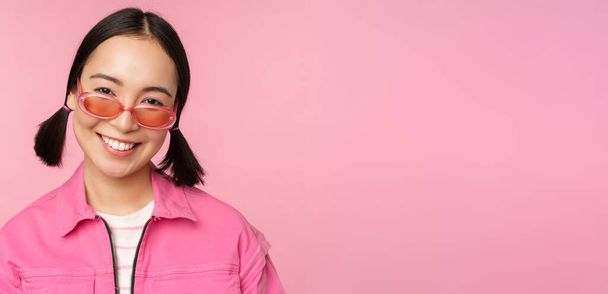 Gros plan de fille coréenne élégante dans des lunettes de soleil, souriant heureux, posant sur fond rose. Les gens face concept - Photo, image