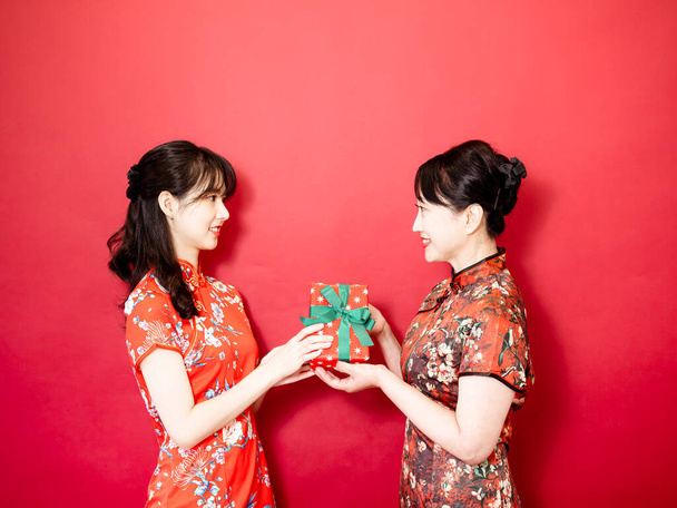 Daughter wearing traditional cheongsam qipao is congratulating mom and giving present wrapped in gift box. Family holiday and togetherness. - Zdjęcie, obraz