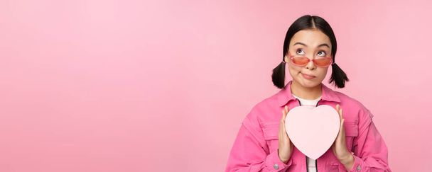 Schöne asiatische Mädchen lächelt glücklich, zeigt Herz Geschenkbox und schaut aufgeregt in die Kamera, steht über rosa romantischen Hintergrund - Foto, Bild