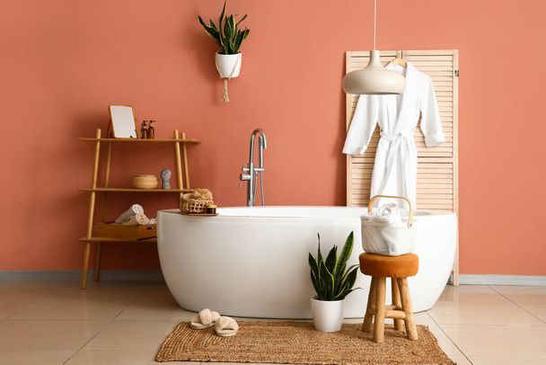 Bañera moderna, plantas de interior, toallas limpias y bata cerca de la pared marrón - Foto, imagen