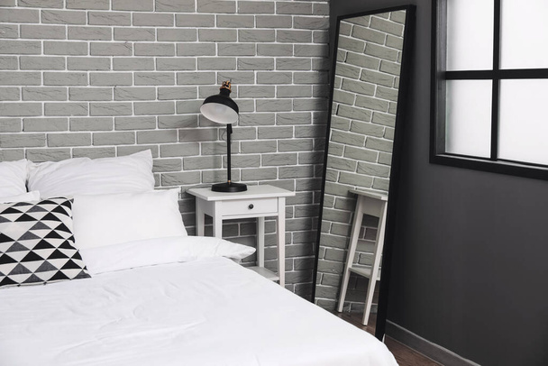 Comfortabel bed, spiegel en moderne lamp op nachtkastje in kamer - Foto, afbeelding