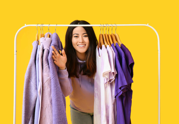 Mujer asiática joven y perchero con ropa en tonos púrpura sobre fondo amarillo - Foto, imagen