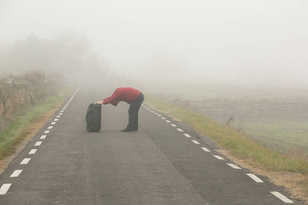 Несучи валізу на руках невпізнаваний чоловік, що йде по дорозі в туманний день
 - Фото, зображення