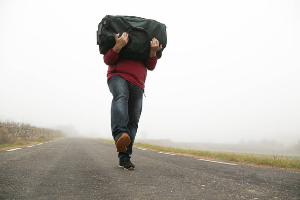 Carregando uma mala em seus braços um homem irreconhecível andando ao longo de uma estrada em um dia nebuloso - Foto, Imagem