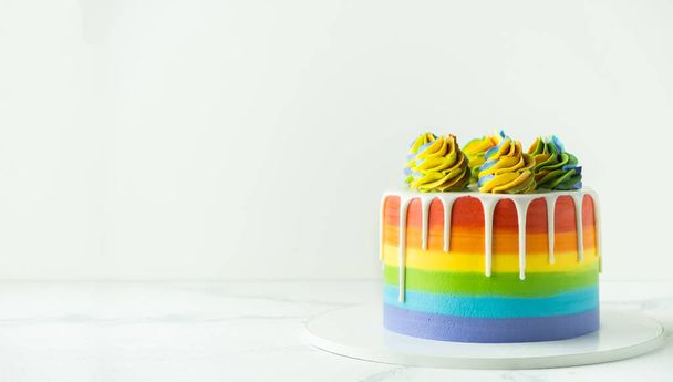 Rainbow cake with whipped cream top on the white background. Birthday cake with multicolored cream cheese frosting and white chocolate drips. - Photo, image