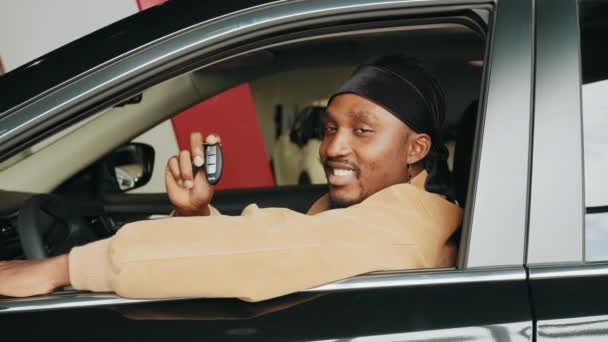 Opgewonden Afrikaan met een autosleutel in zijn nieuwe auto. Op bezoek bij een autodealer. Eigen auto. Vreugdevolle Afro-Amerikaanse man in het bezit van de autosleutel in de auto in de dealer winkel zittend in een nieuwe auto - Video