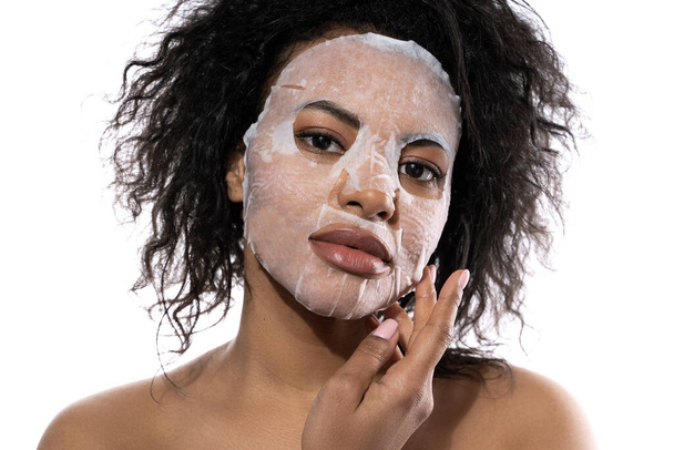 Beautiful black woman with a smooth skin applying facial sheet mask on white background - Fotografie, Obrázek