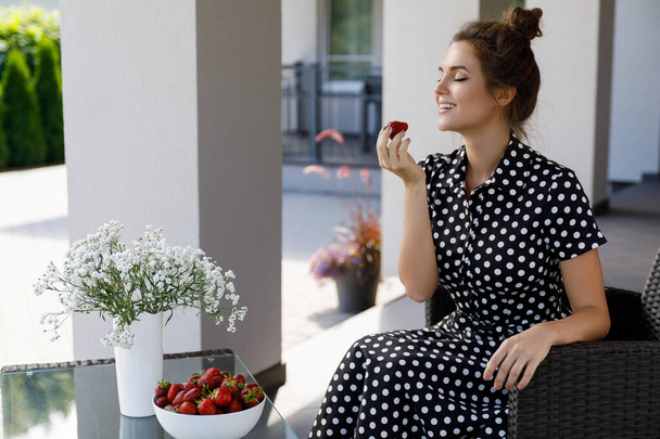 Portrait of gorgeous woman wearing beautiful dress with a polka dot pattern sitting in a patio and eating strawberry - Foto, imagen