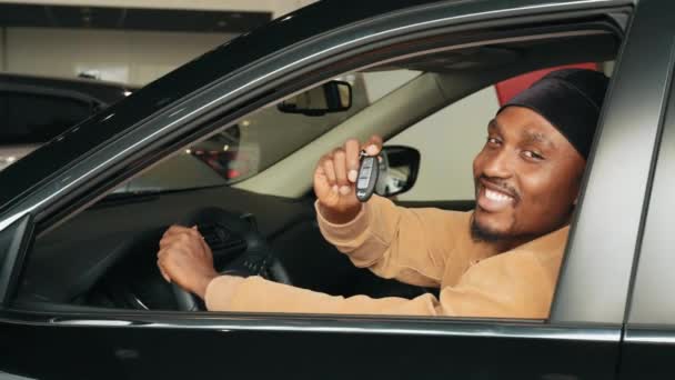 Eigen auto. Opgewonden Afrikaan met een autosleutel in zijn nieuwe auto. Op bezoek bij een autodealer. Vreugdevolle Afro-Amerikaanse man in het bezit van de autosleutel in de auto in de dealer winkel zittend in een nieuwe auto - Video