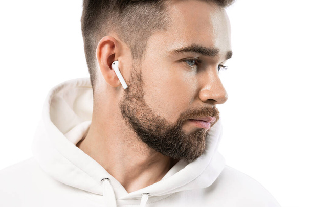 Retrato de hombre barbudo guapo en ropa blanca usando auriculares inalámbricos sobre fondo blanco - Foto, Imagen