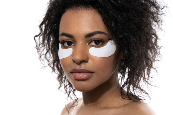 Beautiful black woman with a smooth skin applying adhesive under-eyes patches for dark circles on white background - Valokuva, kuva