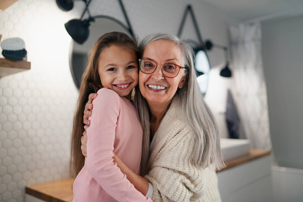 Senior grandmother and granddaughter standing indoors in bathroom, hugging and looking at camera. - Фото, изображение