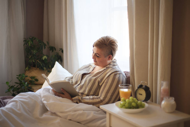 Happy overweight woman reading book in bed at home. - 写真・画像