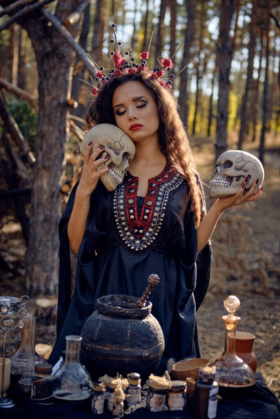 Witch in black, long dress, with red crown in her long, curly hair. Posing in pine forest. Holding skulls. Spells, magic and witchcraft. Close-up. - Zdjęcie, obraz