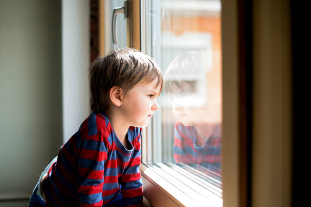 Kid boy sitting near window and looking outside. Serious child attentively looks out of the window - Foto, Bild