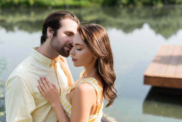 young woman with closed eyes hugging with bearded man near lake - Photo, Image