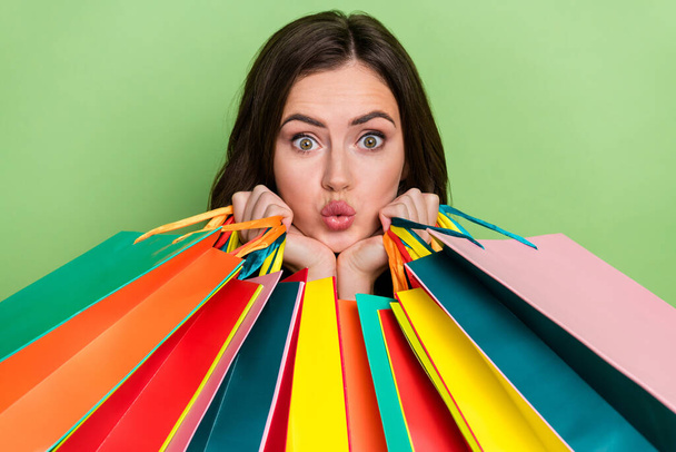 Portrait of attractive stunned wavy-haired girl holding colorful bags pout lips gifts isolated over bright green color background - 写真・画像