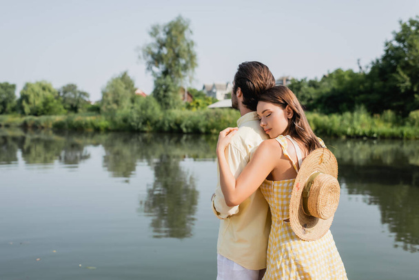 young woman with straw hat hugging boyfriend near lake - Zdjęcie, obraz