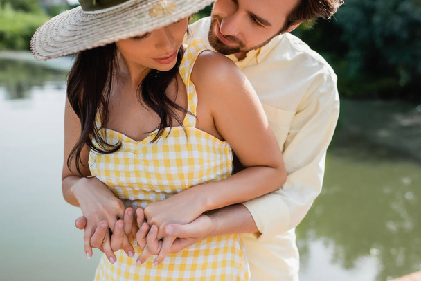 hombre barbudo abrazándose y tomándose de la mano con su novia en sombrero de paja cerca del lago - Foto, imagen
