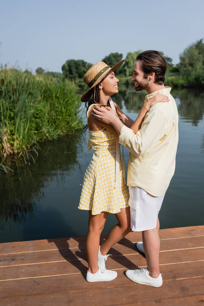 full length of happy young couple in summer clothes hugging on pier near lake - Fotoğraf, Görsel
