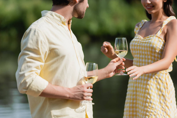 oříznutý pohled na šťastný mladý pár v letním oblečení drží sklenice s vínem v blízkosti jezera - Fotografie, Obrázek