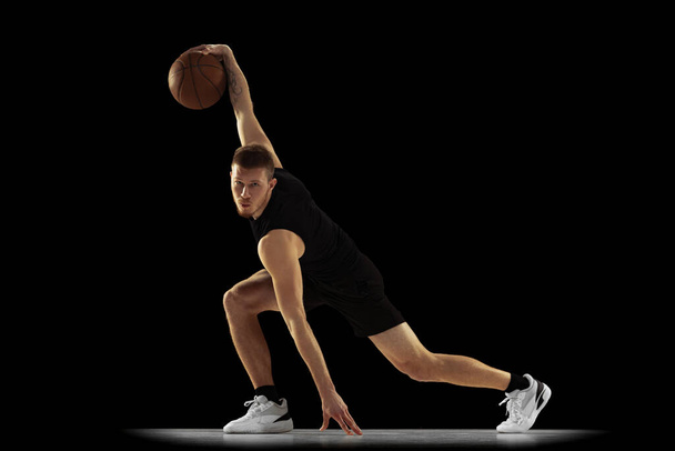 Ground move. Dynamic portrait of young man, basketball player in black uniform training isolated on dark background. Achievements, sport career, motion concepts. - Photo, image