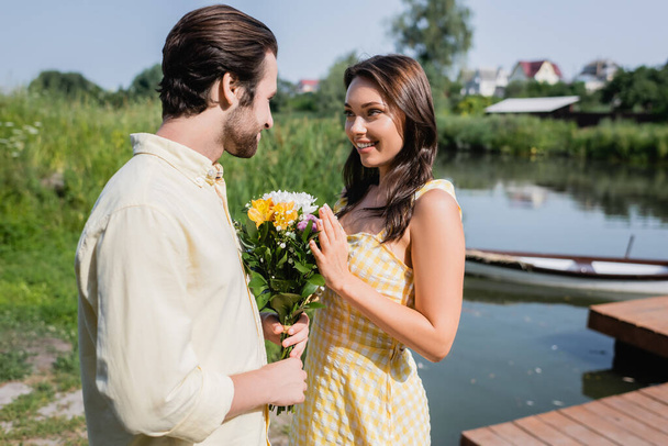 bearded man holding bouquet of flowers near happy woman in dress near lake  - Zdjęcie, obraz