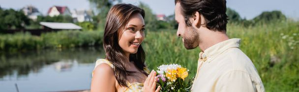 brodaty mężczyzna gospodarstwa bukiet kwiatów w pobliżu szczęśliwy kobieta w sukience w pobliżu jeziora, sztandar - Zdjęcie, obraz