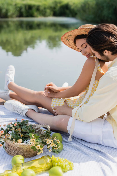 romantic couple sitting and holding hands on pier near lake during picnic - Photo, image