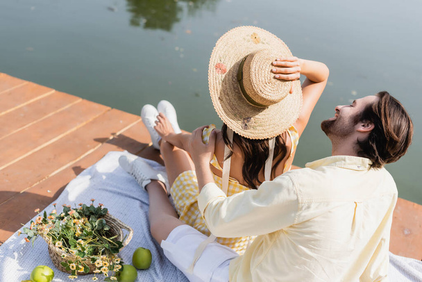 high angle view of cheerful man looking at girlfriend in straw hat while sitting on pier during picnic - Φωτογραφία, εικόνα