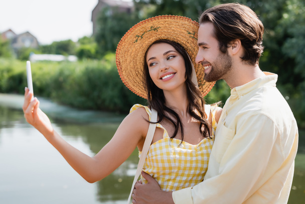 feliz joven mujer en sombrero de paja tomando selfie con novio cerca del lago - Foto, imagen