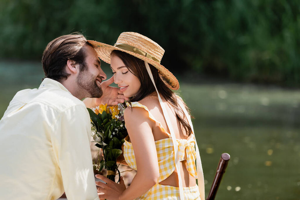 happy young woman in straw hat holding bouquet of flowers near romantic boyfriend on lake - Photo, Image