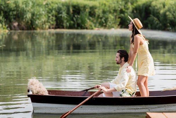 ロマンチックなボート旅行中に男の後ろに立つ藁帽子の幸せな若い女性の側面図  - 写真・画像