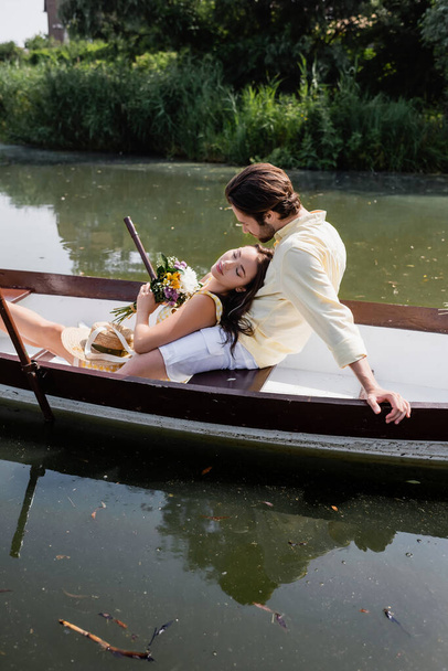 young woman with closed eyes holding bouquet of flowers and lying in boat with man  - Foto, Imagem