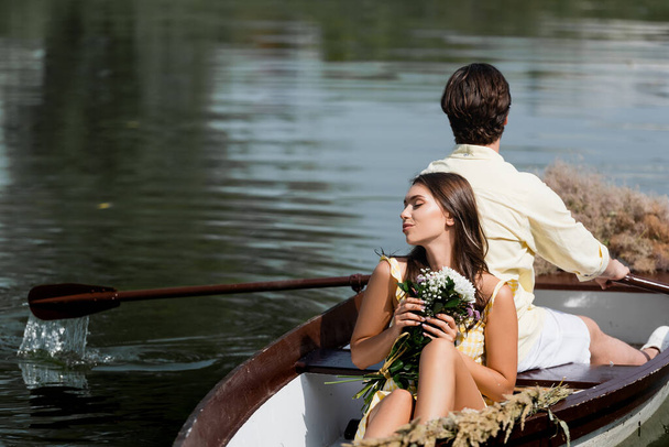 молодая женщина с цветами и опираясь на спину человека во время романтической поездки на лодке  - Фото, изображение