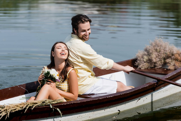 happy young woman holding flowers and leaning on back of boyfriend during romantic boat trip  - Fotoğraf, Görsel