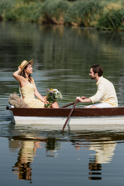 side view of smiling woman in straw hat holding flowers and having romantic boat ride with man  - Photo, Image