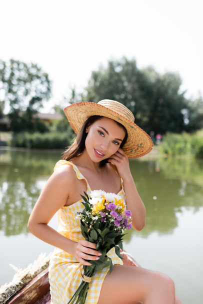 pretty young woman in straw hat and dress holding flowers while sitting near lake - Fotografie, Obrázek