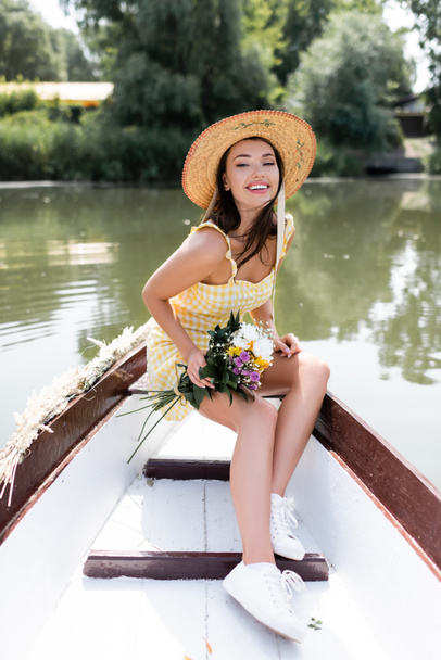 joyeuse jeune femme en chapeau de paille et robe tenant des fleurs tout en ayant promenade en bateau sur le lac - Photo, image