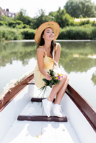 pozytywna młoda kobieta w słomkowym kapeluszu trzyma kwiaty podczas przejażdżki łodzią po jeziorze - Zdjęcie, obraz