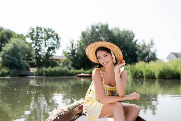 χαμογελαστή νεαρή γυναίκα με ψάθινο καπέλο και φόρεμα που έχει βάρκα βόλτα στη λίμνη - Φωτογραφία, εικόνα