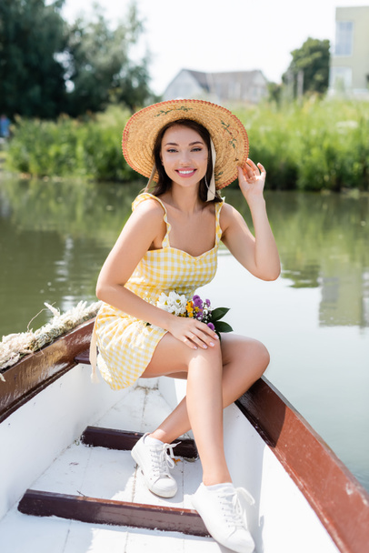 позитивная молодая женщина регулирует соломенную шляпу и катание на лодке по озеру - Фото, изображение