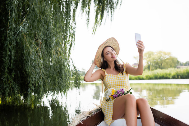 alegre joven mujer en sombrero de paja y vestido tomando selfie durante paseo en barco en el lago - Foto, Imagen