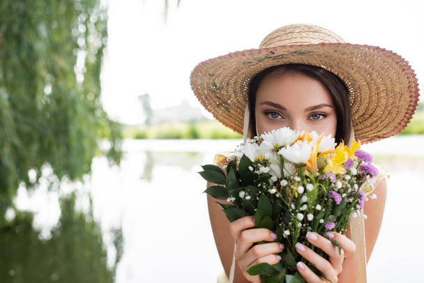 mujer joven en sombrero de paja cubriendo la cara mientras huele flores  - Foto, Imagen