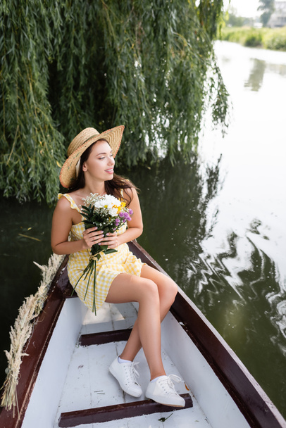 jeune femme heureuse en chapeau de paille et robe tenant des fleurs pendant la promenade en bateau sur la rivière - Photo, image